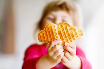 Portrait of happy little preschool girl holding fresh baked heart waffle. Smiling hungry toddler child with sweet biscuit wafer. Sweet sugar belgian waffles.
